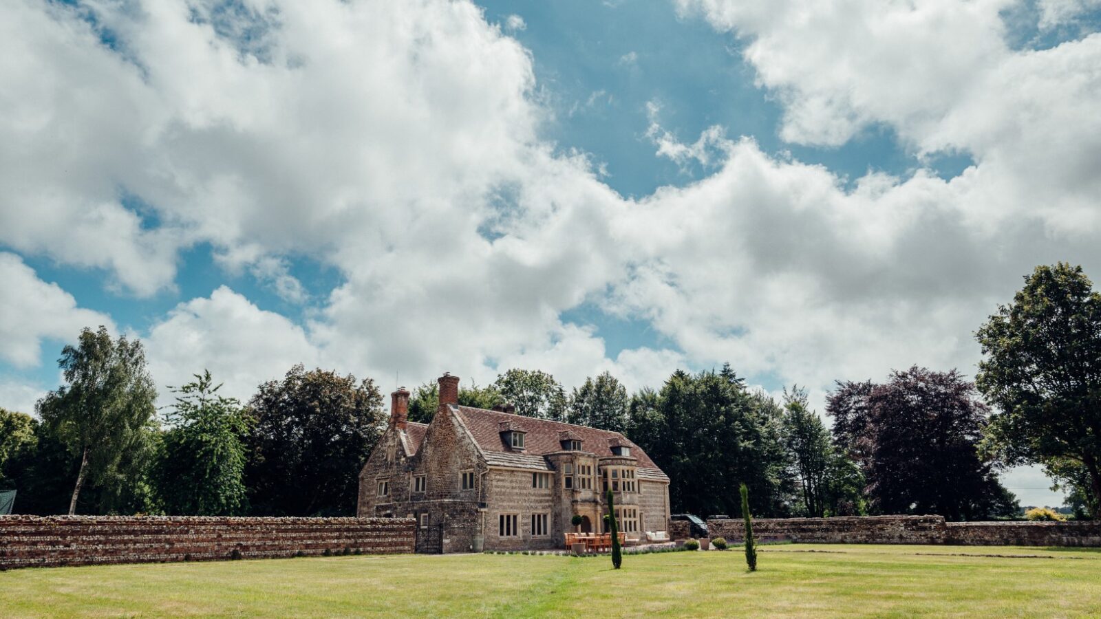 Almer Manor, a stone house with a red-tiled roof, is gracefully nestled amidst a grassy field and trees under a partly cloudy blue sky.