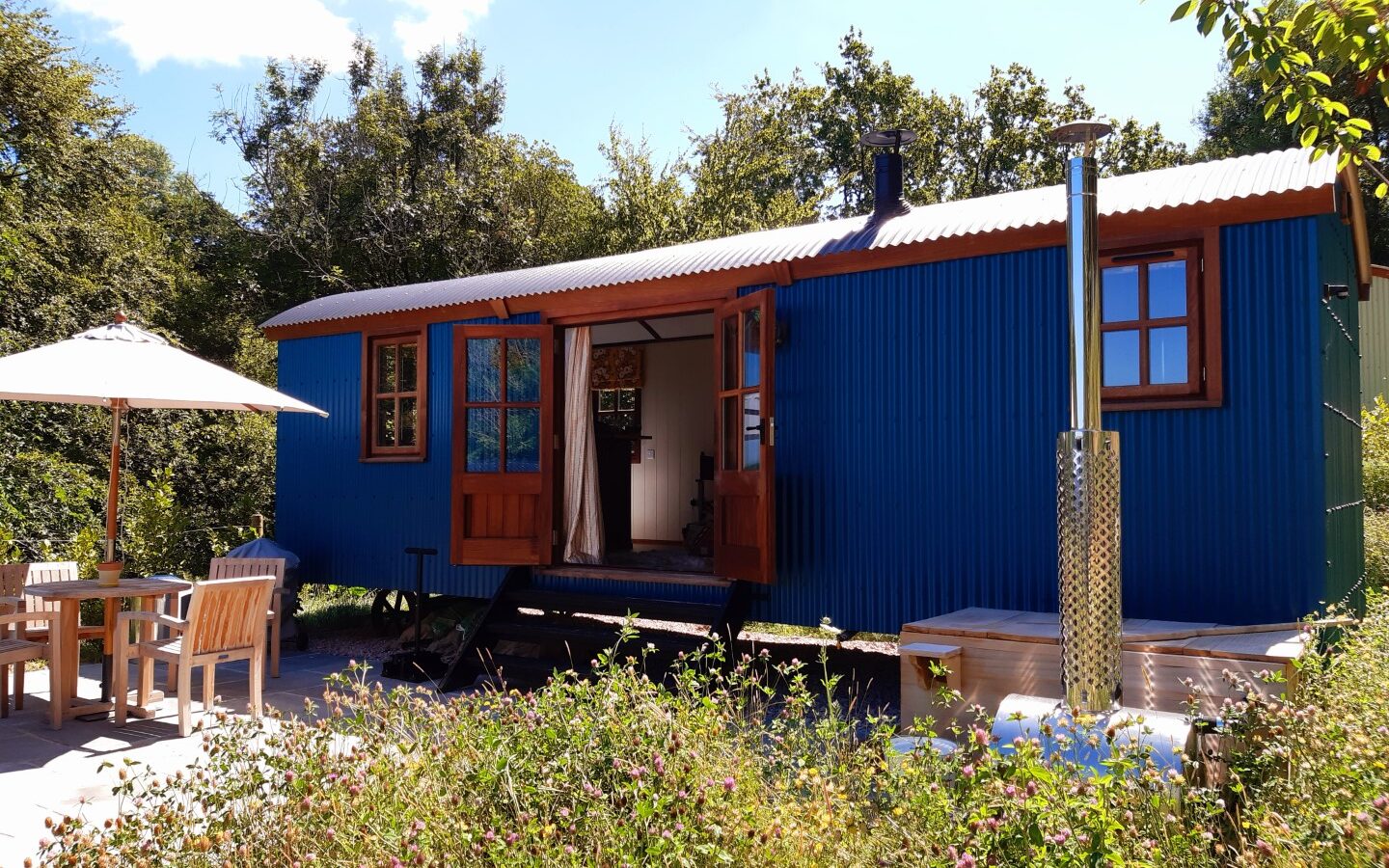 A cozy, vibrant blue tiny house with wooden accents and open doors sits amidst lush greenery at Dittisham Hideaway. A small patio with a table, chairs, and patio umbrella is visible, offering a serene outdoor space under a clear sky reminiscent of Shepherds Lodges.