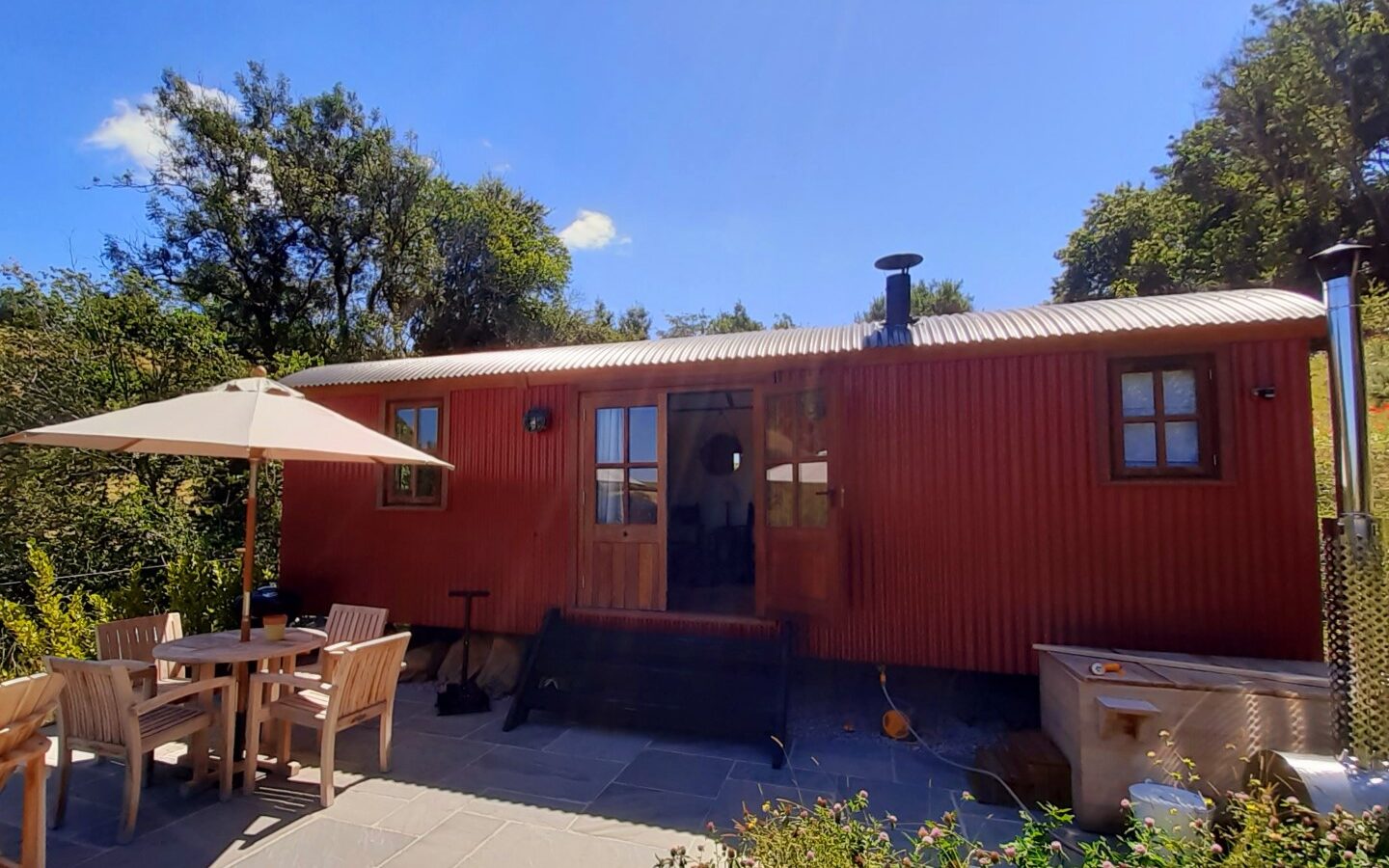 A rustic red cabin, reminiscent of the charming Shepherds Lodges at Dittisham Hideaway, boasts wooden doors and windows, nestled among trees. A sunny patio with a table, chairs, and an umbrella completes this serene setting.