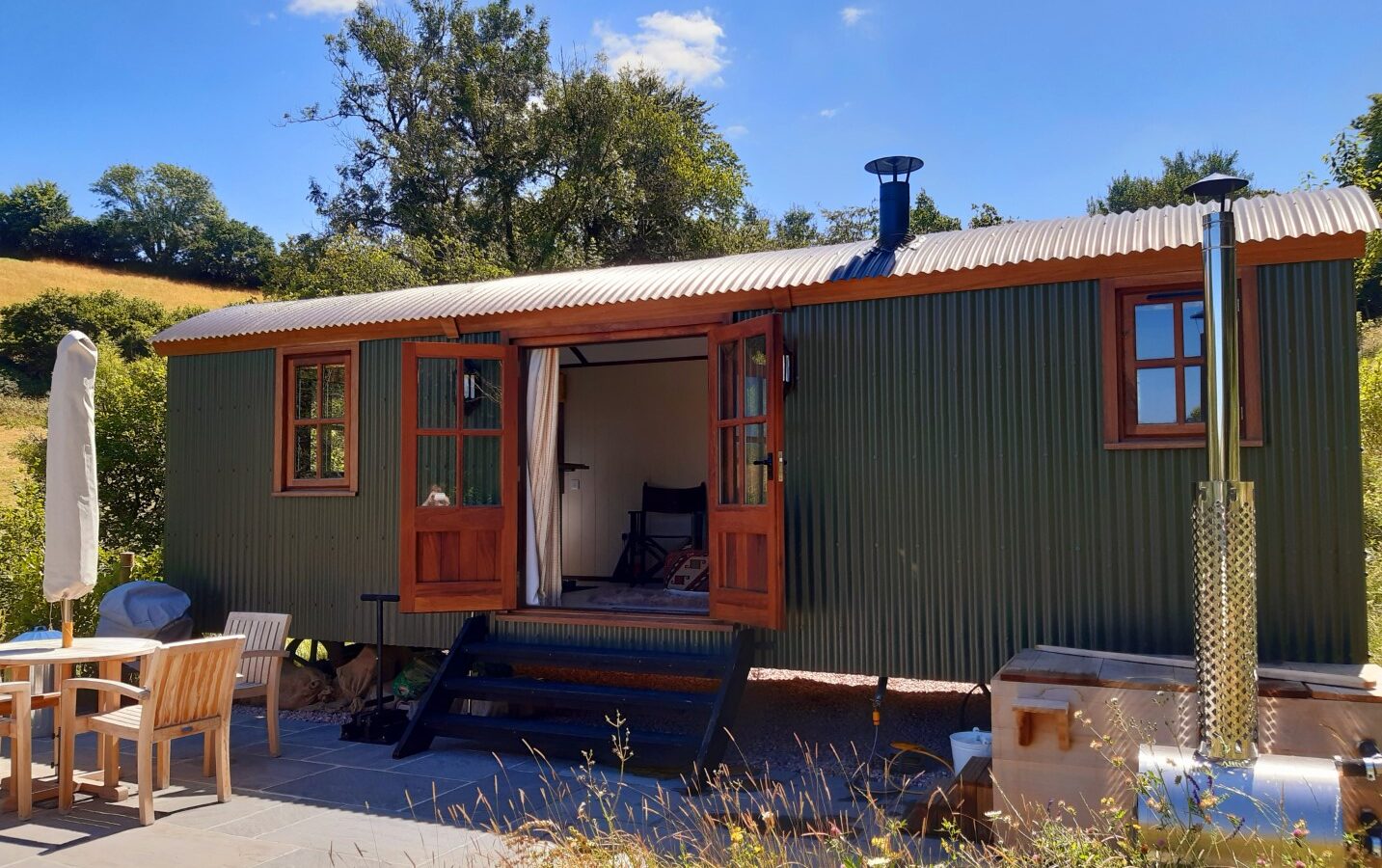 Nestled in the serene countryside, this Dittisham Hideaway features a green, rustic tiny house with wooden trim and open doors. Amidst lush greenery under a clear blue sky, it offers patio chairs, a table, and a wood-fired hot tub. Tall trees frame this idyllic Shepherds Lodge escape.