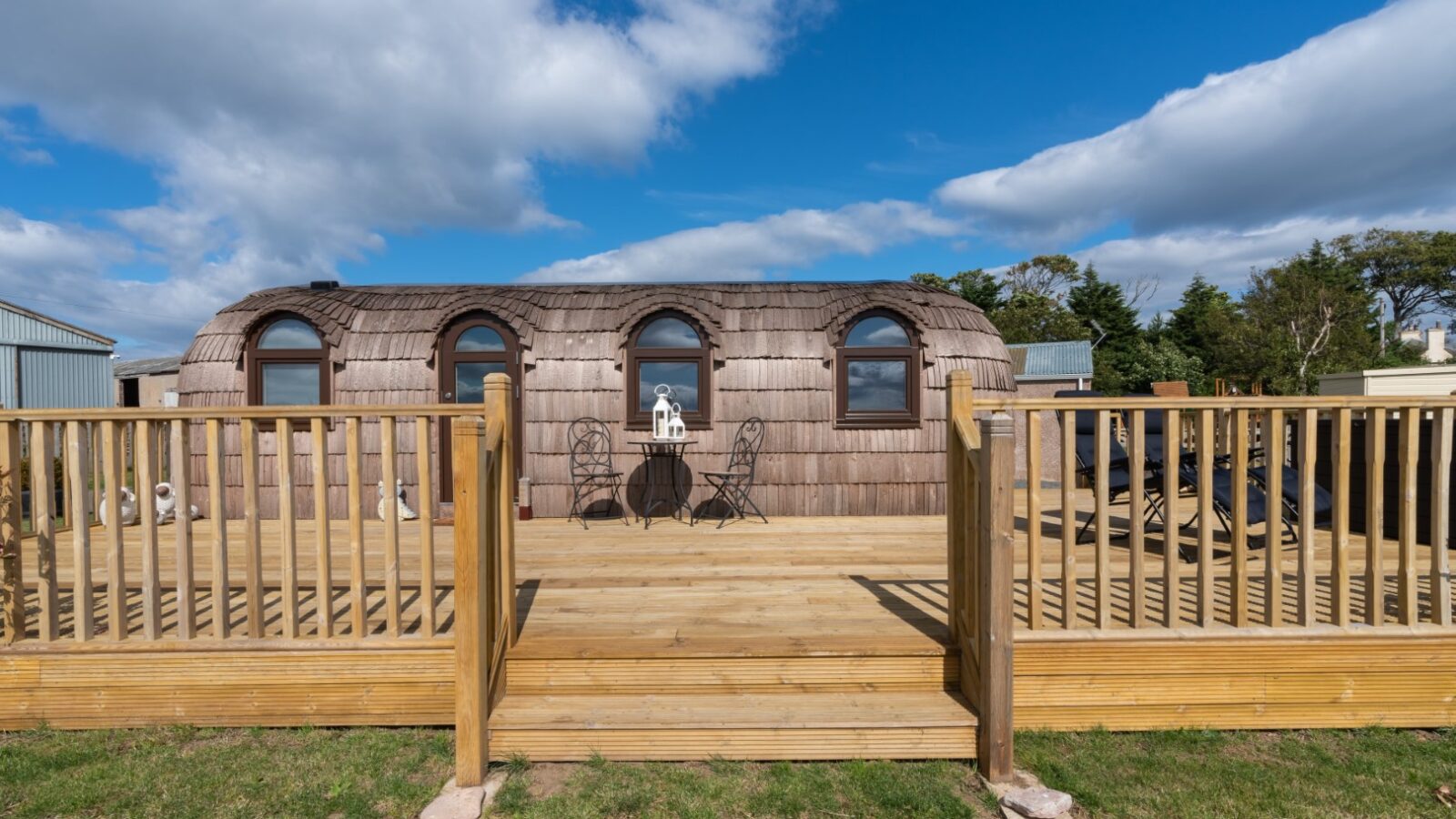 This wooden cabin, reminiscent of a castle with its arched windows and shingled roof, sits on a deck complete with railing and patio furniture, perfect for serene escapes under a clear blue sky.