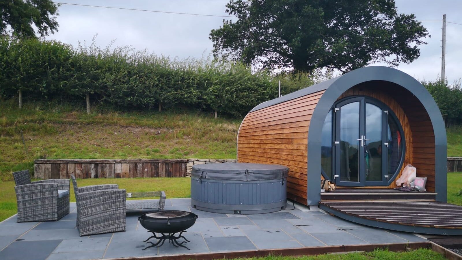 Experience glamping at its finest in our wooden pod cabin with glass doors. Nestled on the picturesque Great House Farm, it features a hot tub and outdoor seating on a patio, surrounded by lush grass and trees beneath a cloudy sky.