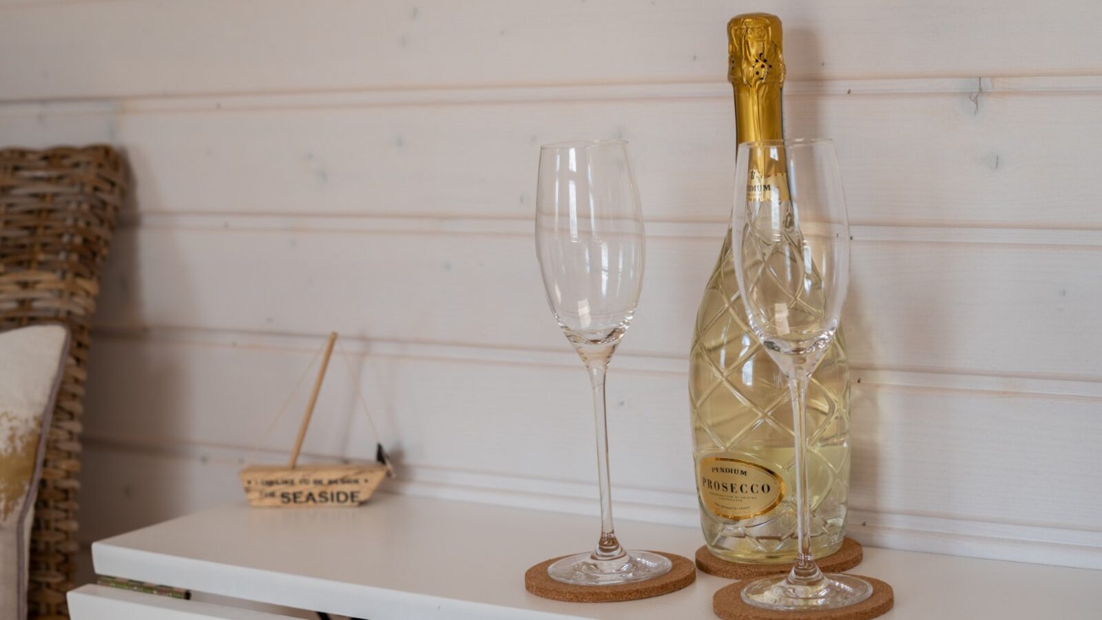 Two empty wine glasses and a bottle of prosecco rest on coasters atop a white surface, with a small sign reading 