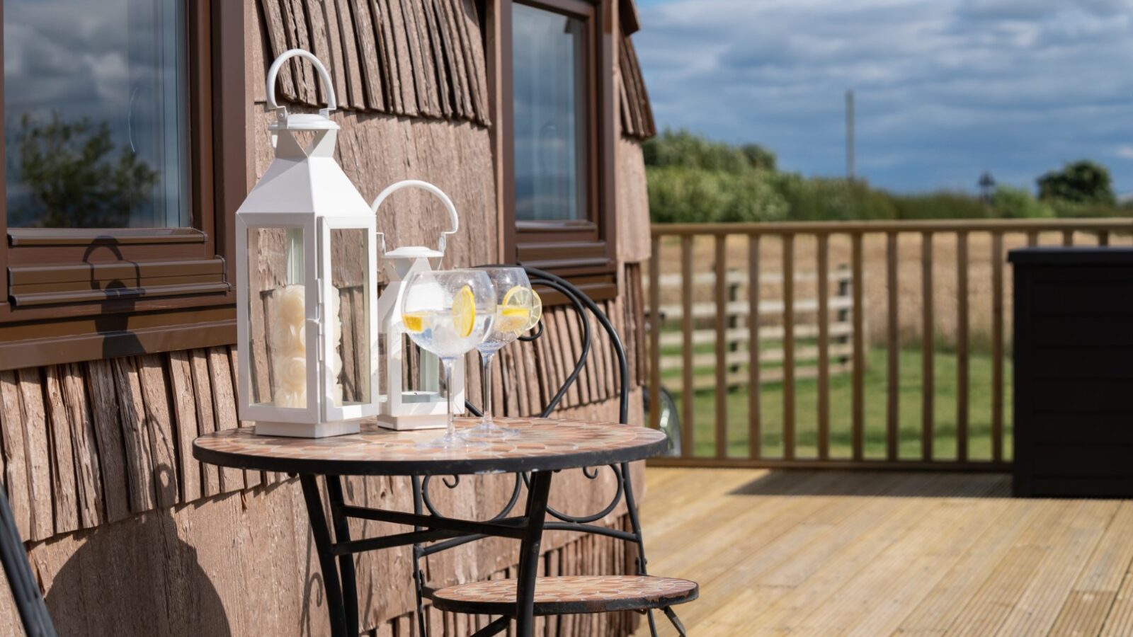 Outdoor patio escape with a small round table, two lanterns, and three glasses filled with drinks. Wooden decking and lush greenery provide a serene backdrop reminiscent of a secluded rock & castle setting.