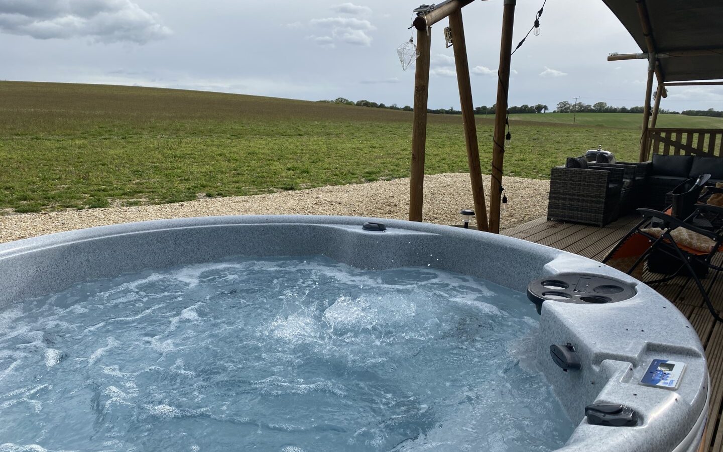 An outdoor hot tub with bubbling water sits on a wooden deck overlooking the vast Chilterns under a cloudy sky. Nearby, seating under a safari tent offers a cozy retreat.