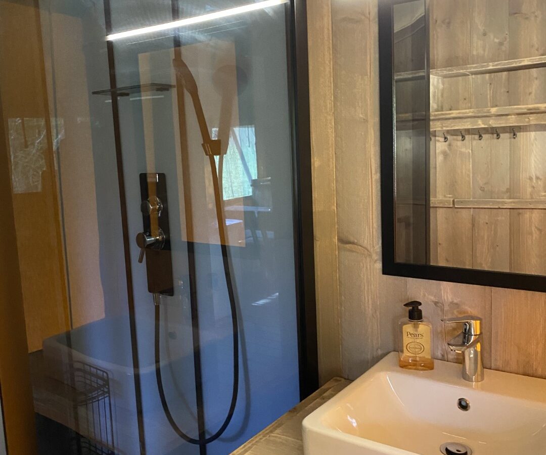 This modern bathroom exudes a Chilterns charm with its glass shower enclosure, wooden walls, square sink on a wooden counter, and a rectangular mirror adorned with lights. It's the perfect blend of contemporary design and the rustic allure of a Safari Tent experience at Chilterns View.