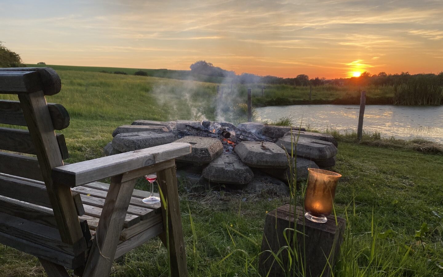 A wooden chair and candle sit by a smoldering fire pit near a pond at the Chilterns View Safari Tent, with the sun setting over grassy fields in the background.