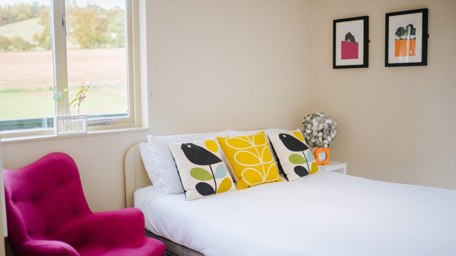 The Wills Treehouse bedroom features a bright ambiance with a double bed, pink chair, colorful pillows, and two framed pictures on the wall. Enjoy the serene window view of fields that brings nature right into your cozy retreat.