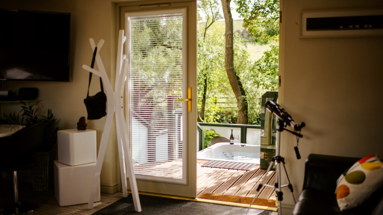 Room with a glass door opens to a deck at Wills Treehouse, featuring a hot tub and telescope, surrounded by trees and lush greenery in the background.