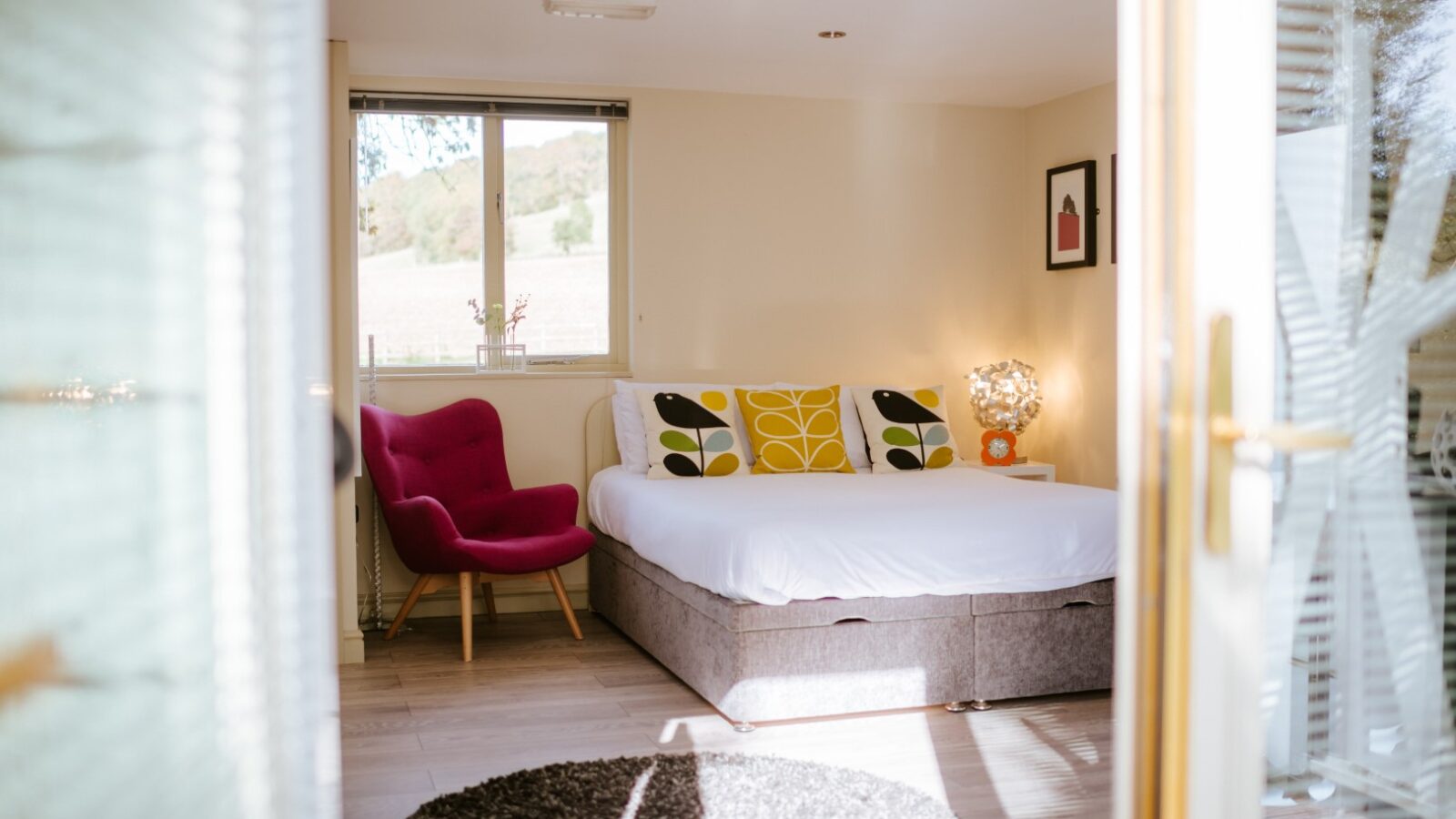 A bedroom with a double bed, colorful pillows, a pink chair, and a round rug offers a tranquil retreat. The window frames an enchanting view reminiscent of Wills Treehouse's leafy embrace outside.