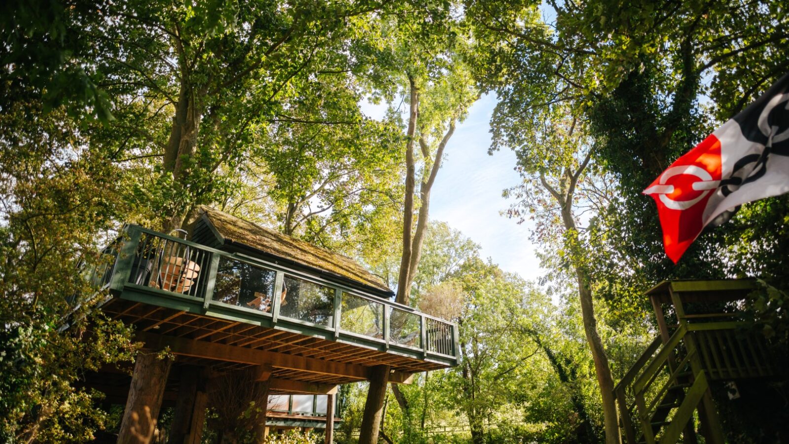 Wills Treehouse, surrounded by lush greenery, boasts a wooden deck and a colorful flag on the right.