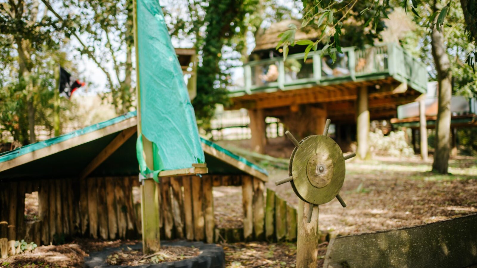 Nestled in a wooded area, the wooden play structure boasts a green sail and steering wheel, evoking the charm of a Chicken Shed Lodge. Elevated treehouses dot the background, creating an adventurous haven for young explorers.