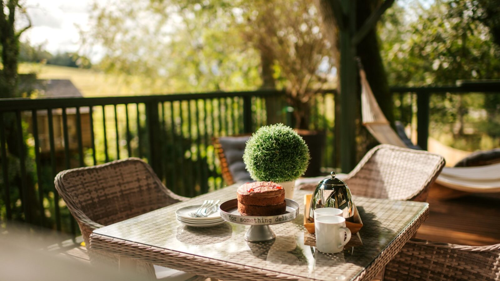 Wicker patio set on a deck with a cake, metal teapot, and cup on the table, surrounded by Wills Treehouse's lush greenery and a hammock swaying gently in the background.