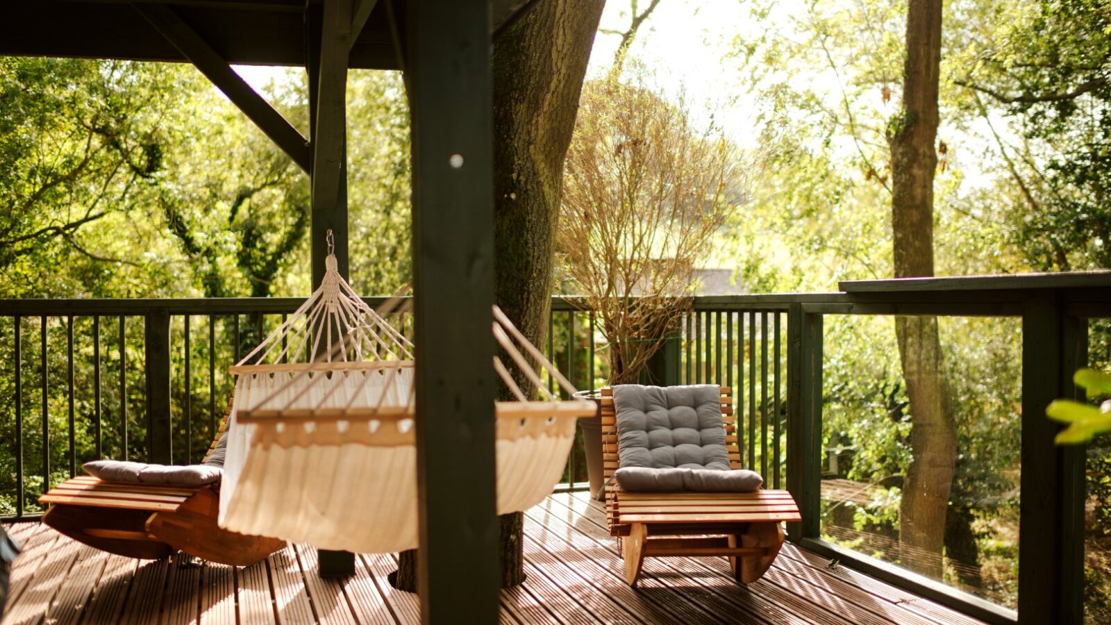 A wooden deck with a hammock and two cushioned lounge chairs, nestled high in Wills Treehouse among the trees and greenery on a sunny day.