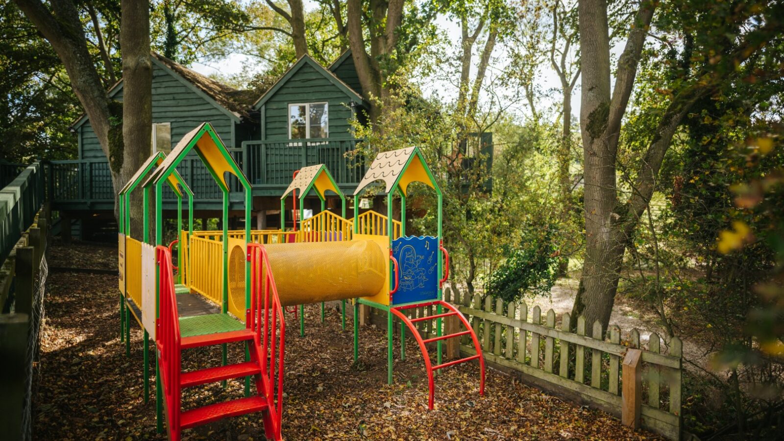 A colorful playground with slides and a tunnel is nestled among the trees, featuring a charming wooden treehouse in the background.