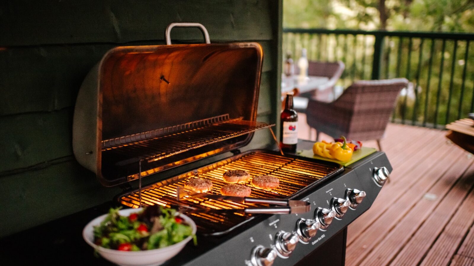 A gas grill with burgers sizzling at Wills Treehouse, a bottle of sauce ready to add flavor, peppers on the side, and a fresh salad bowl nearby on the rustic wooden deck.