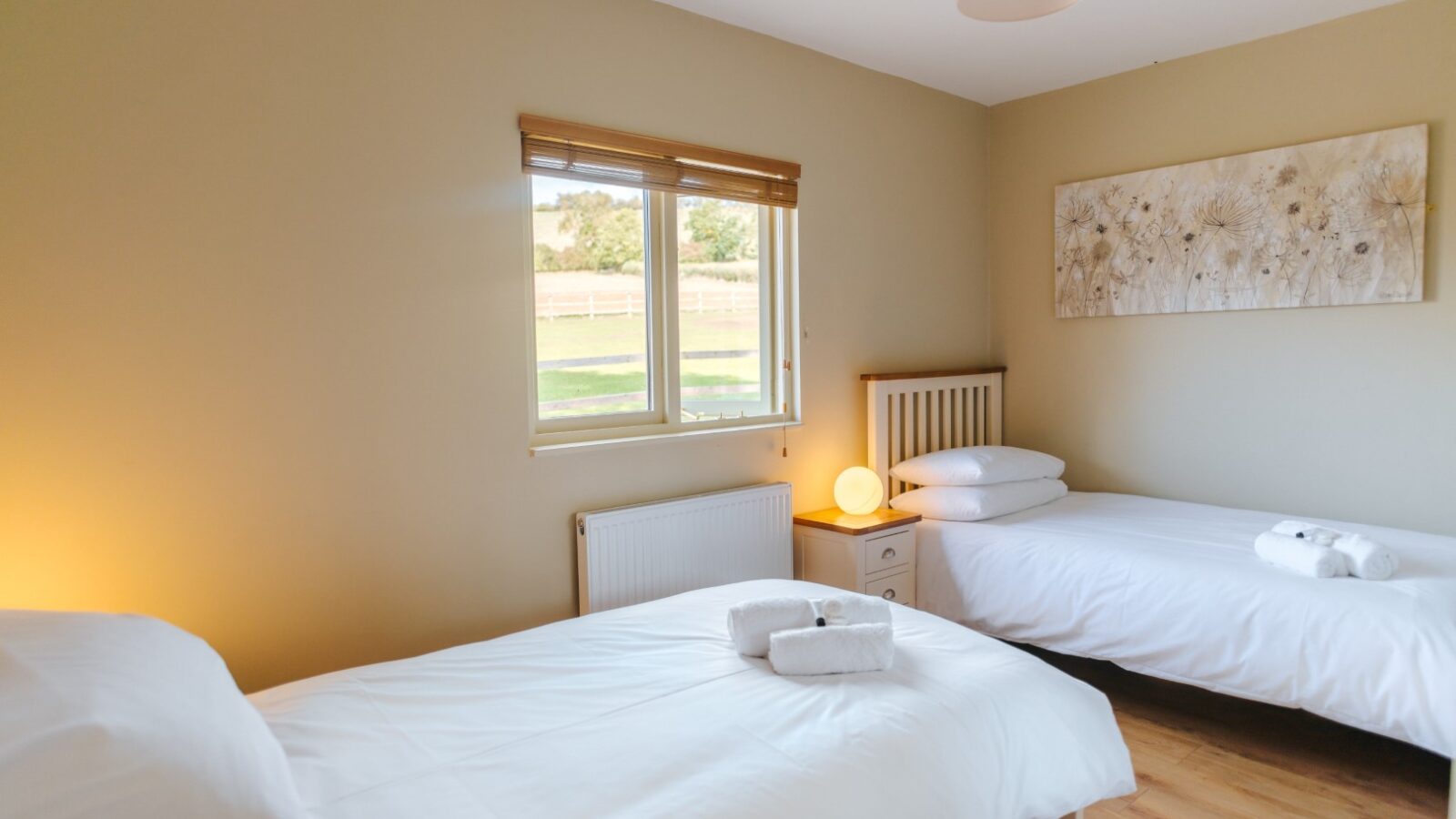 A minimalist bedroom at Chicken Shed Lodge features two single beds, white bedding, a nightstand, and a window offering tranquil views of the greenery outside.