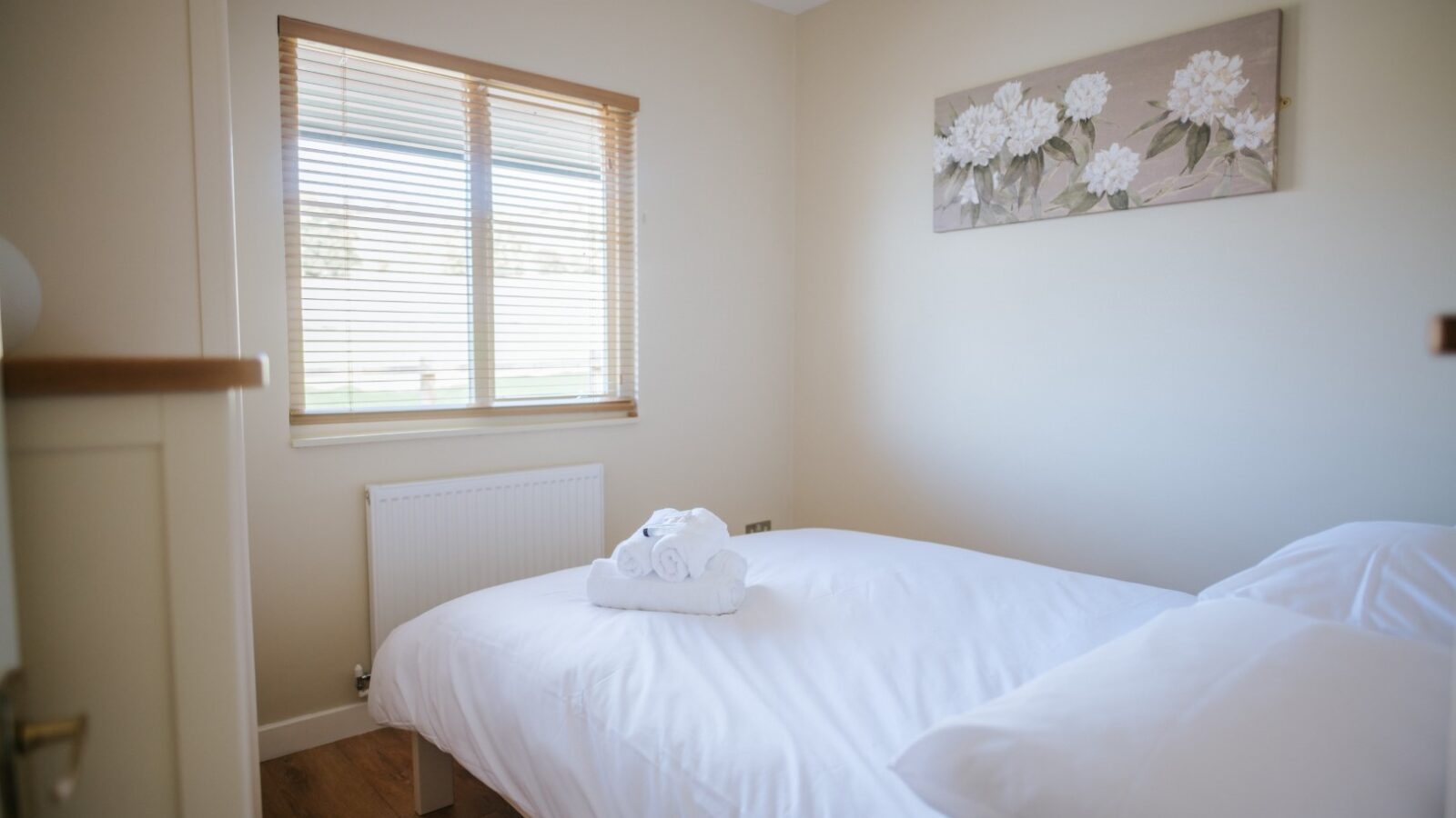 The Chicken Shed Lodge offers a cozy and tidy bedroom featuring a neatly made bed, folded towels, a window with blinds, and a floral picture on the wall.