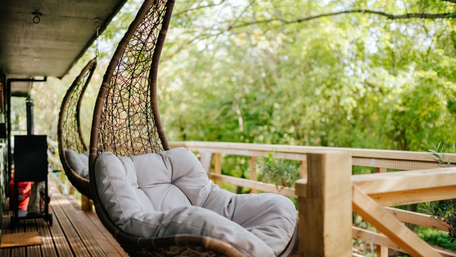 Wicker hanging chairs with cushions on the wooden deck of Chicken Shed Lodge overlook a lush, green forest.