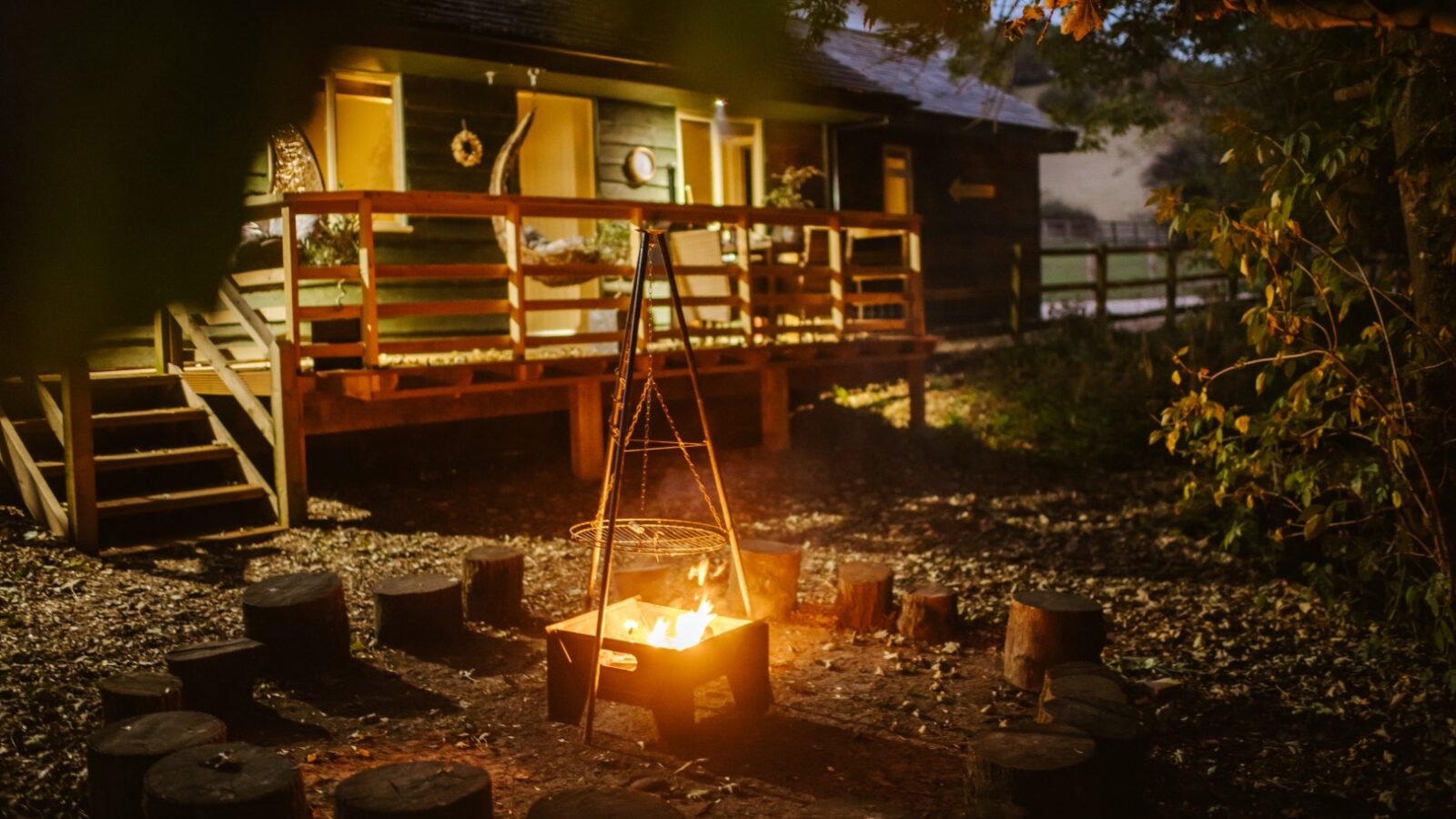 The Chicken Shed Lodge, a cozy cabin with a porch, is illuminated by a campfire surrounded by wooden stumps for seating, nestled in a wooded area at dusk.