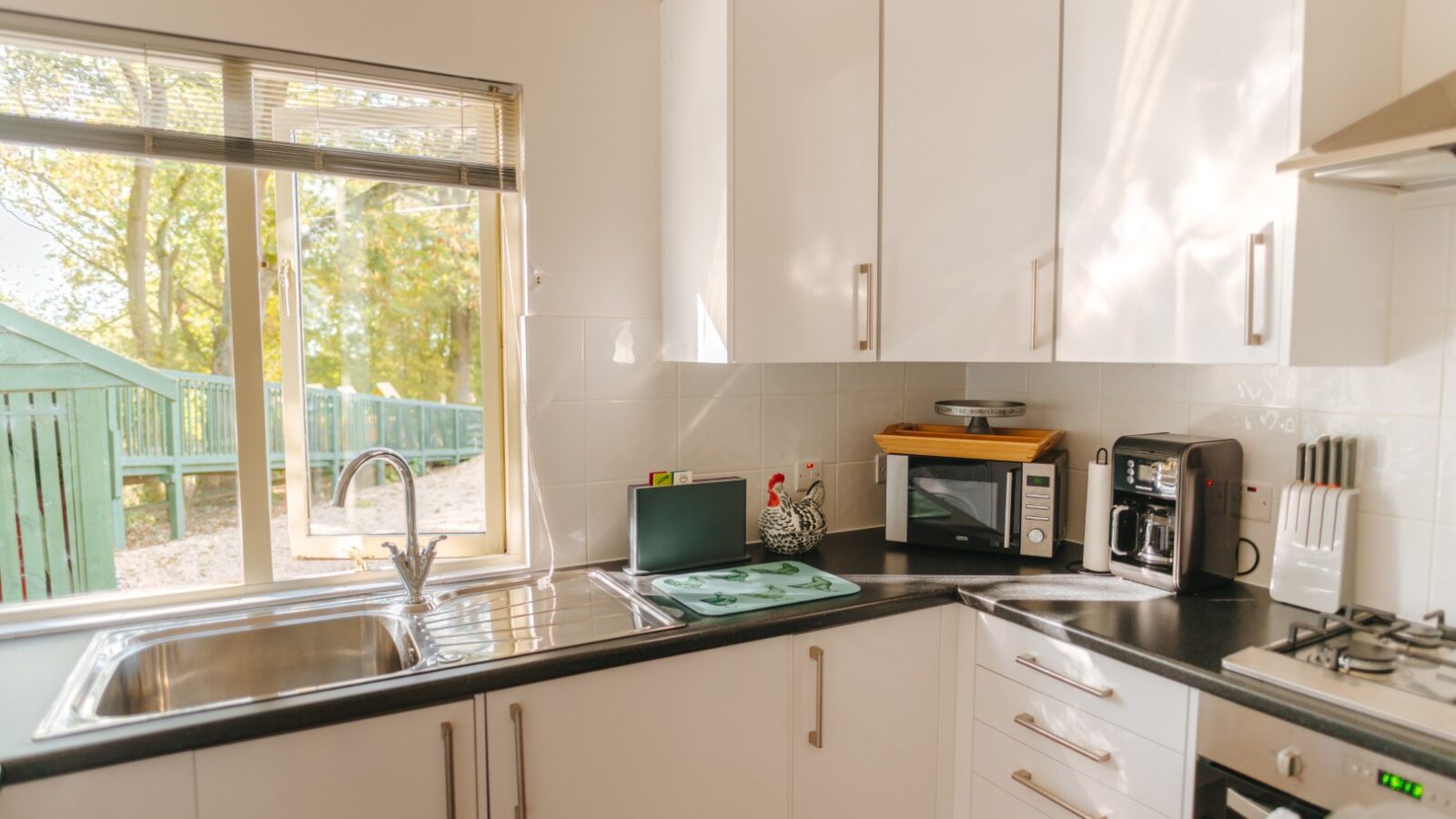 Modern kitchen at Chicken Shed Lodge with white cabinets, black countertop, stainless steel sink, microwave, and coffee maker, all beautifully illuminated by natural light.