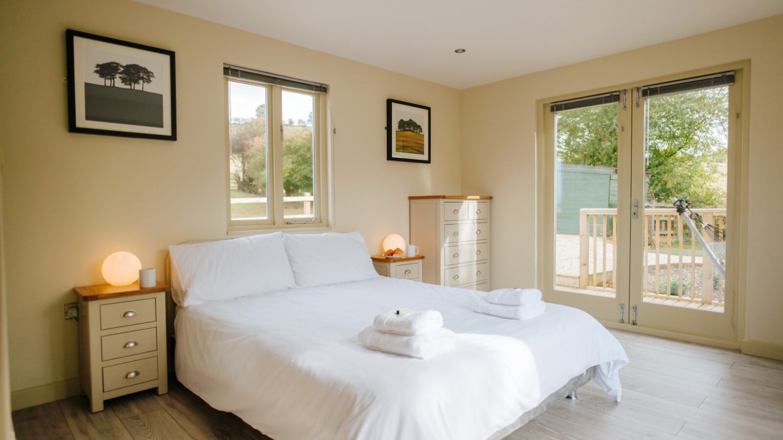 A bedroom at Bob's Lodge features a neatly made white bed, two bedside tables with lamps, artwork adorning the walls, and large windows offering a stunning view.