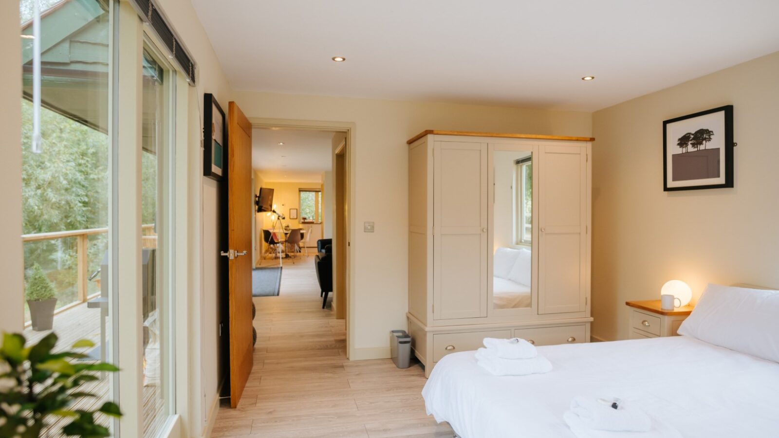 Bright bedroom at Bob's Lodge, featuring a large window, double bed, wardrobe, and an open door leading to a cozy living area. Fresh towels are neatly placed on the bed.