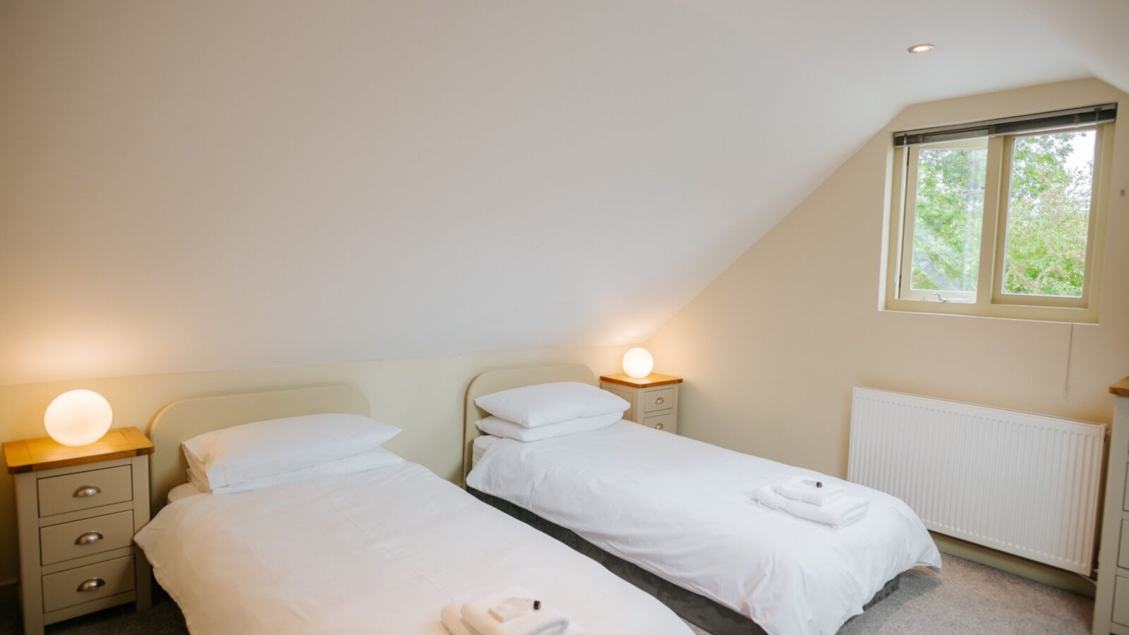 The attic bedroom at Bob's Lodge features two single beds, bedside tables with lamps, a window, and towels neatly placed on the beds.