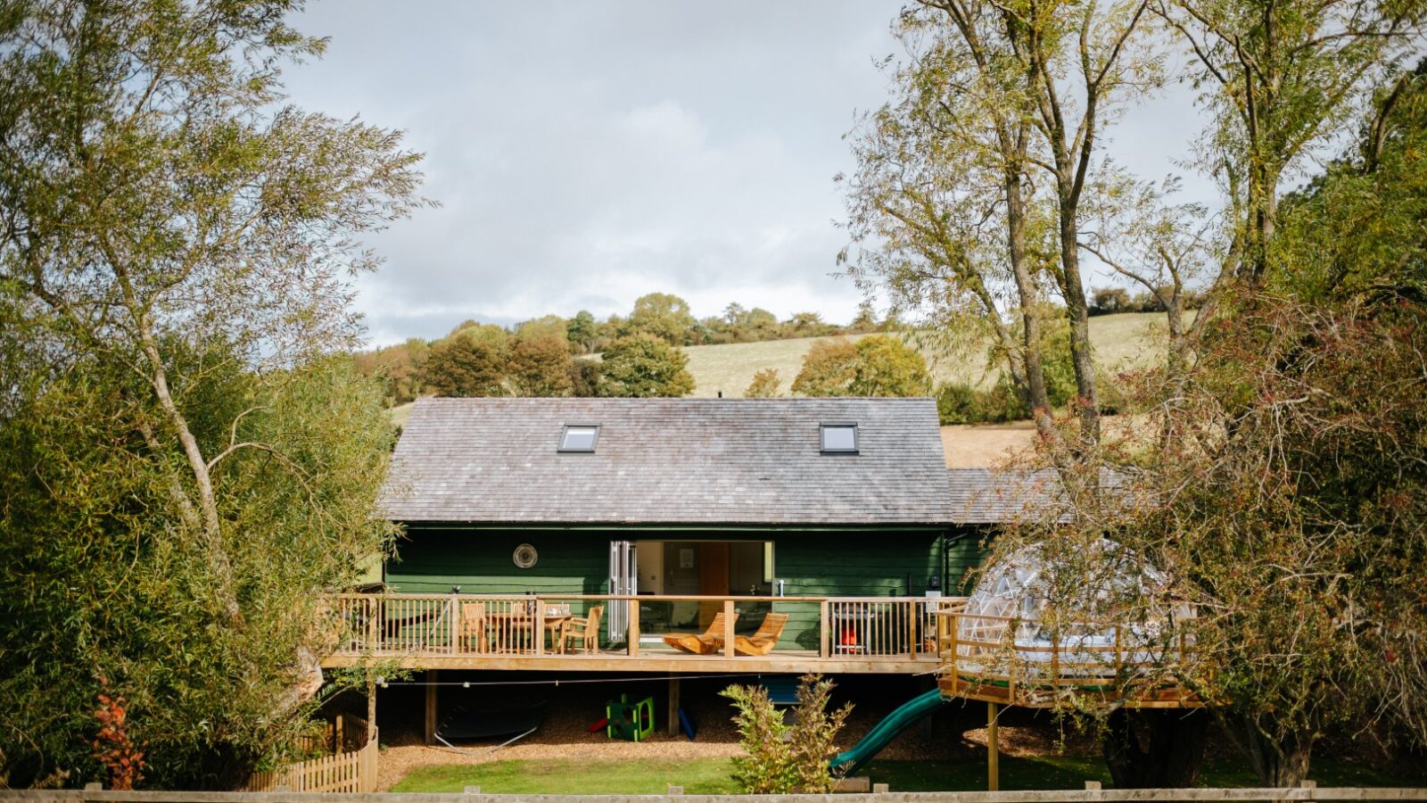 Bobs Lodge, a quaint green cabin with a wooden deck, is nestled in a rural landscape, surrounded by trees and featuring a small fenced yard.