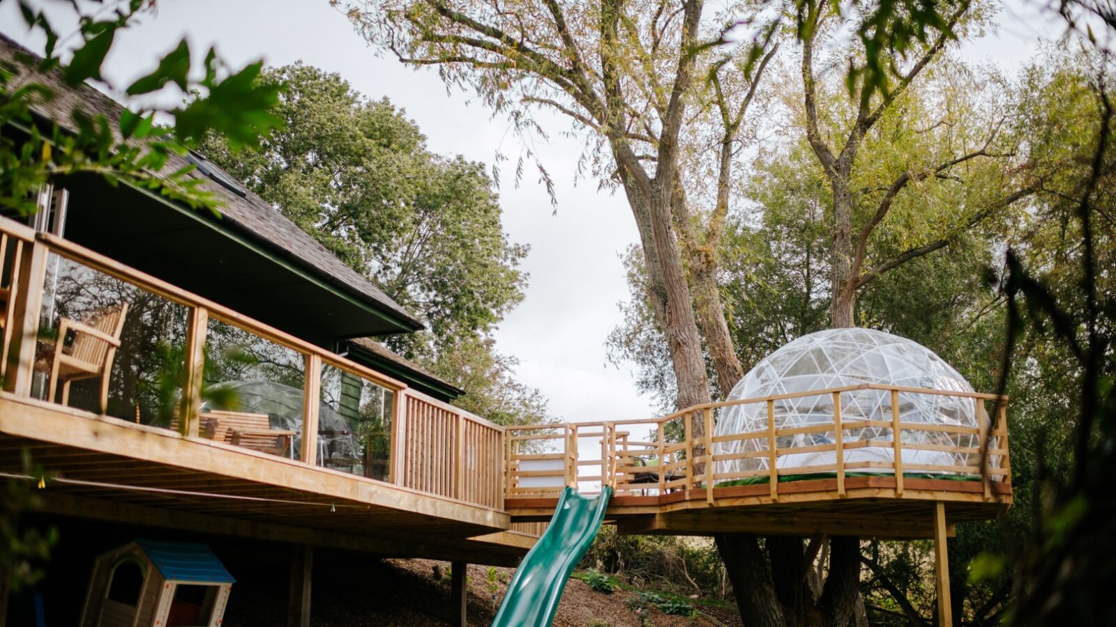 A modern treehouse named Bob's Lodge features a wooden deck, slide, and geodesic dome nestled among the trees, with a small doghouse nearby.