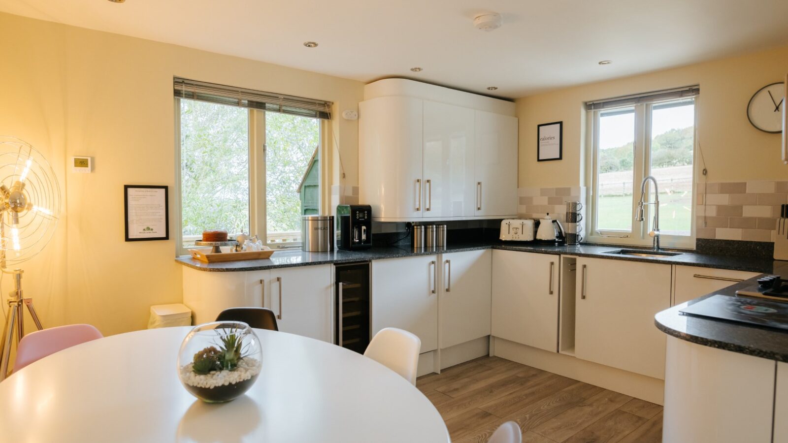 Modern kitchen at Bob's Lodge featuring white cabinets, a round dining table, wooden floors, and large windows overlooking lush greenery.