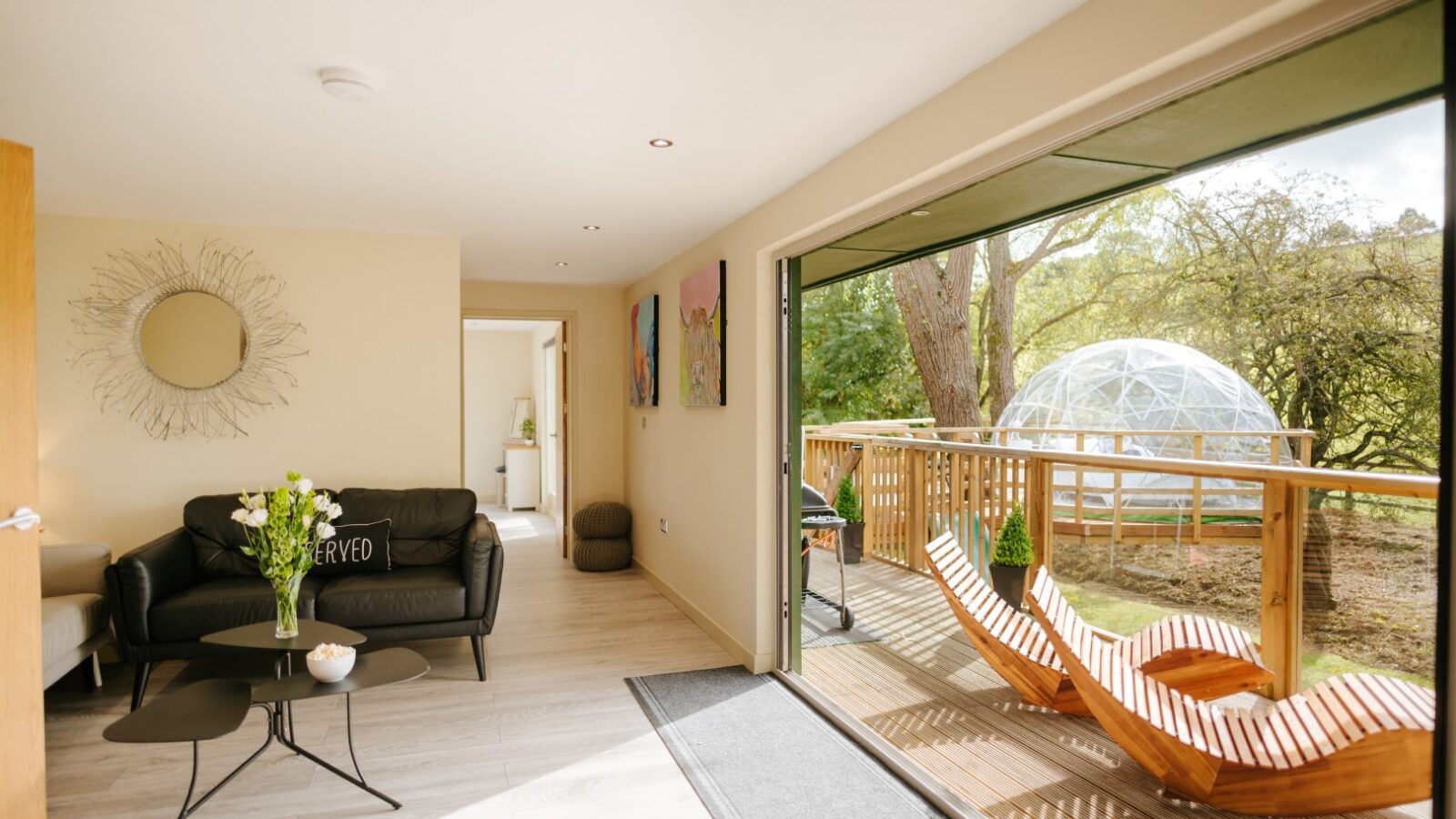 The modern living room at Bob's Lodge features sleek black sofas, a round mirror, and large glass doors that open to a deck with two wooden loungers and a striking geodesic dome.