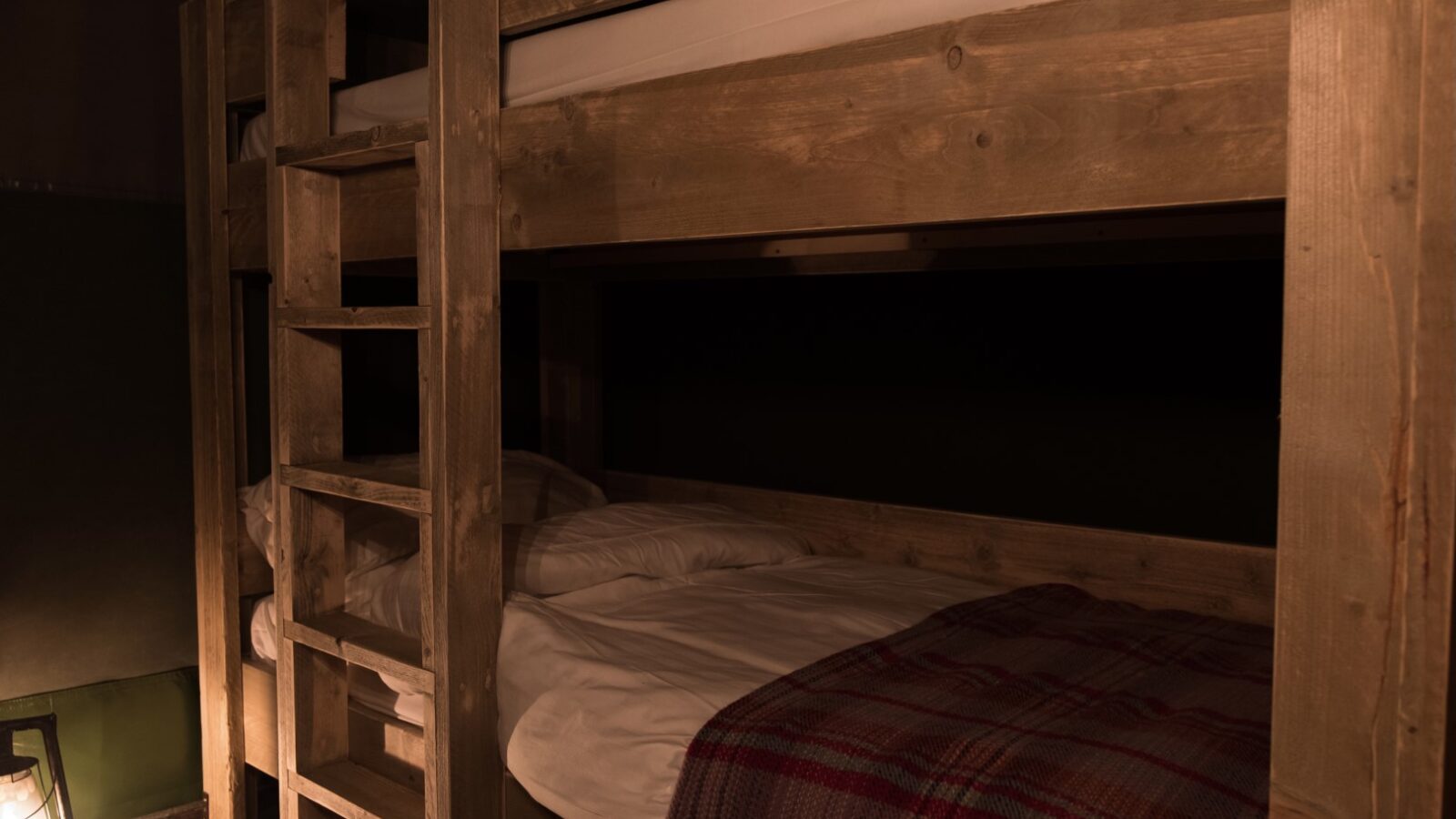 A cozy wooden bunk bed with white bedding and a plaid blanket, softly lit by a lantern on the floor, offers a warm retreat reminiscent of charming lodges at Sloeberry Farm.