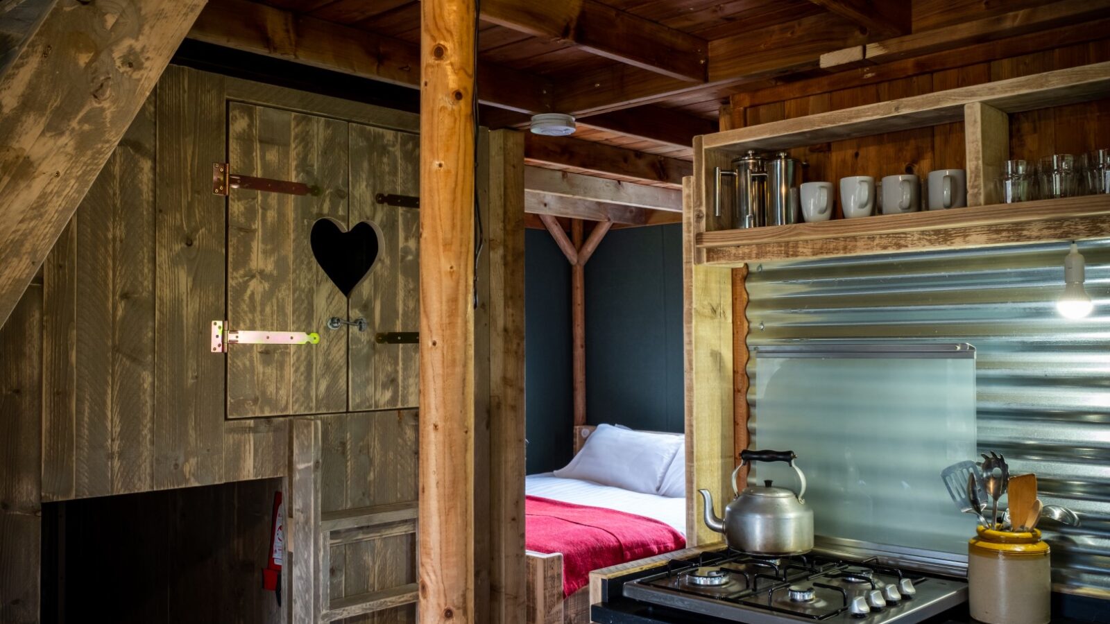 Cozy wooden cabin interior at Sloeberry Farm with a kitchenette, stove, teapot, and bed adorned with a red blanket. Rustic decor elements add charm to your stay in these delightful farm lodges.