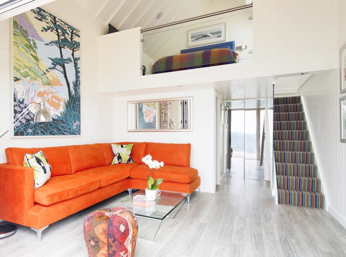 Bright living room with an orange sofa, glass coffee table, colorful artwork, and striped stairs leading to a lofted bedroom area. The design evokes the charm of Cary Arms suites, blending vibrant colors reminiscent of cozy beach huts.