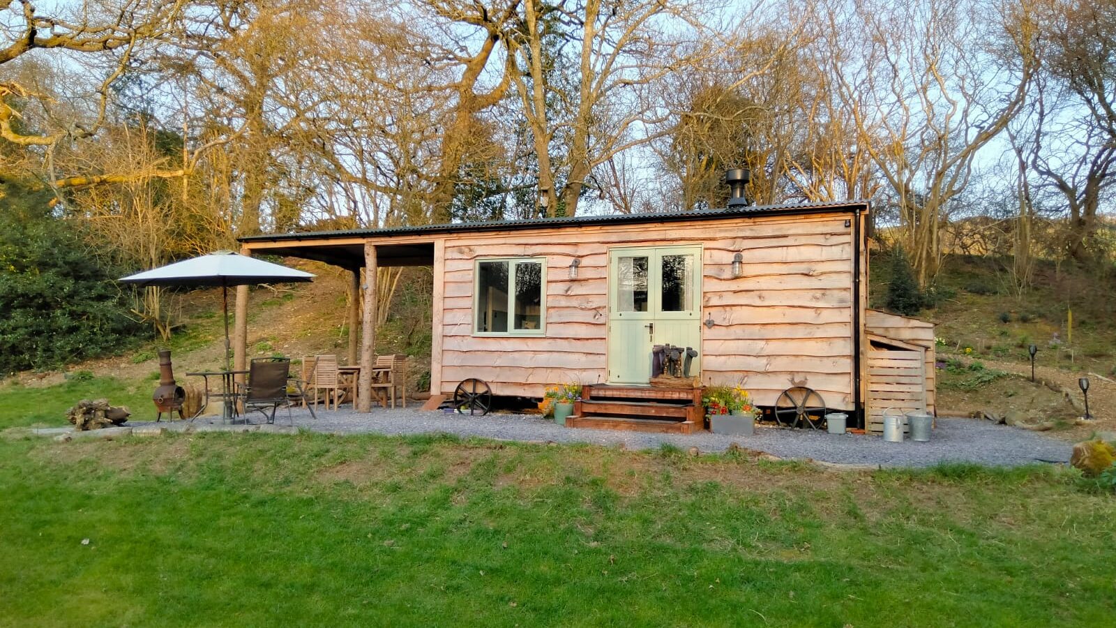 A small wooden cabin, known as Caban Eric, sits in a grassy area surrounded by bare trees. Its patio features chairs and a parasol, inviting guests to relax in nature's tranquility.