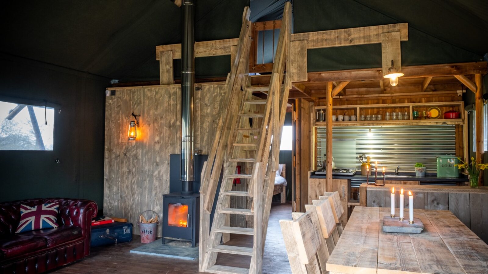 The rustic interior of the lodge at Sloeberry Farm features a wooden ladder, a cozy dining table, a leather sofa, and a wood-burning stove snugly set beneath the pitched roof.