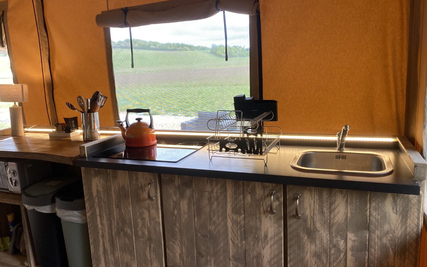 A rustic kitchen setup in the Chilterns View Safari Tent features a sink, dish rack, stove, and kettle. A large window offers a stunning view of the grassy fields outside.