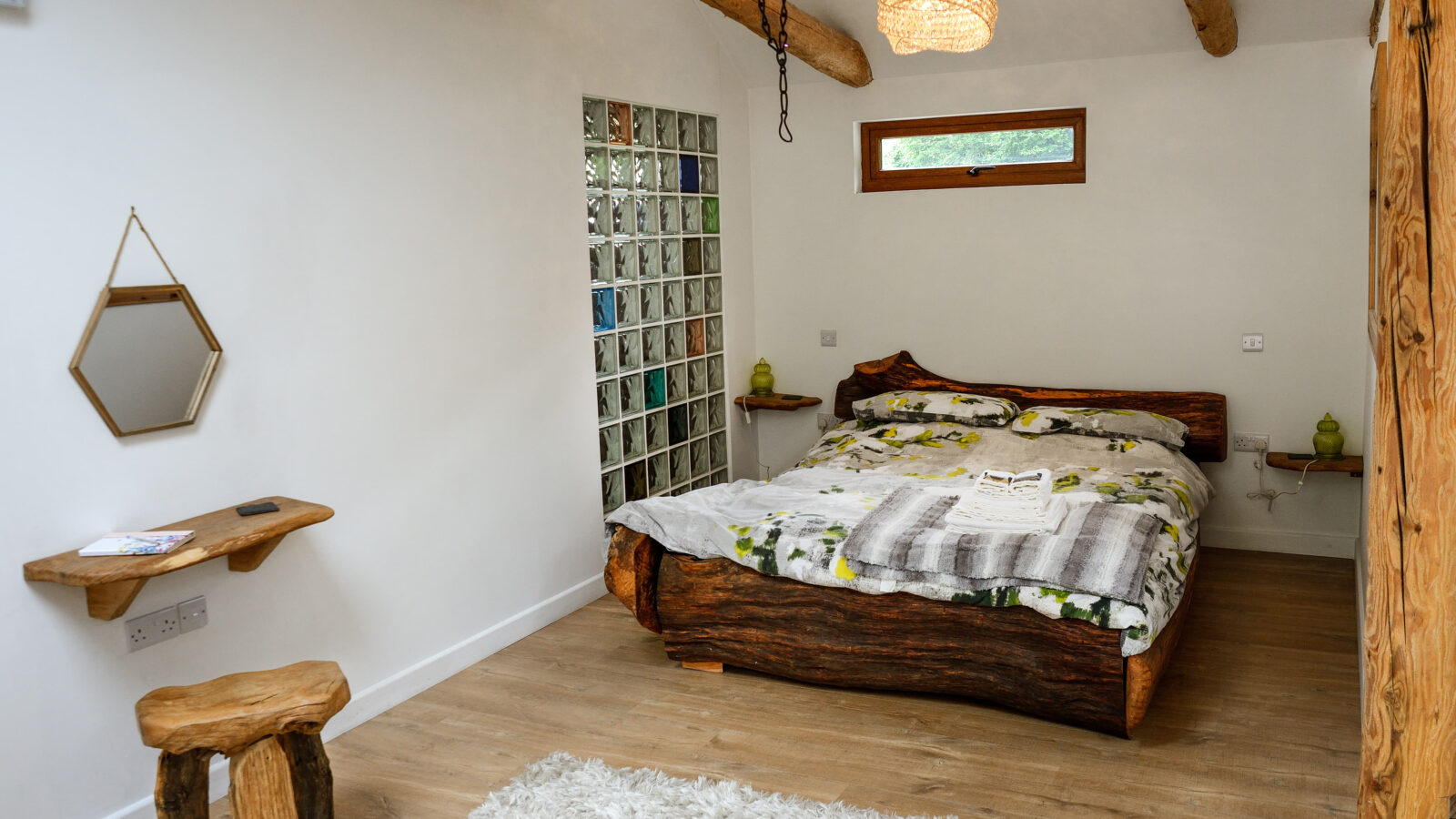 A rustic bedroom at Nant Awen Cottages features a wooden bed, wooden beams, a small window, a wood shelf with a mirror, and a fluffy white rug.