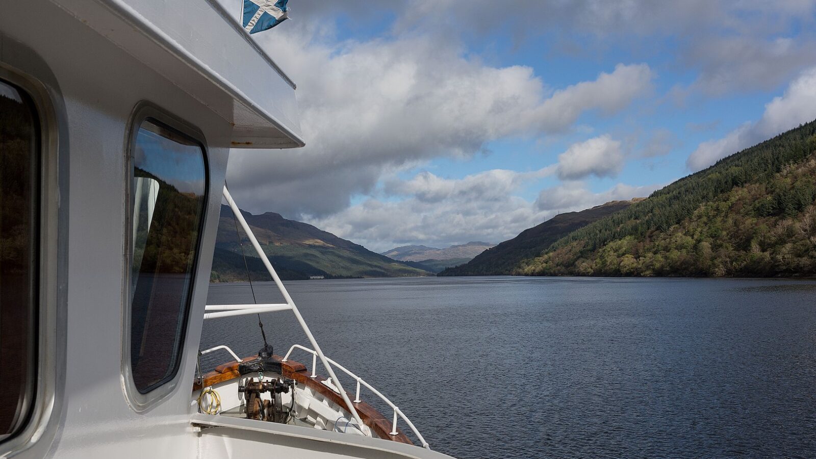 A boat from Argyll Cruising is navigating through a calm lake surrounded by lush, green hills. The sky above is partly cloudy, and a small Scottish flag flutters proudly on the boat.