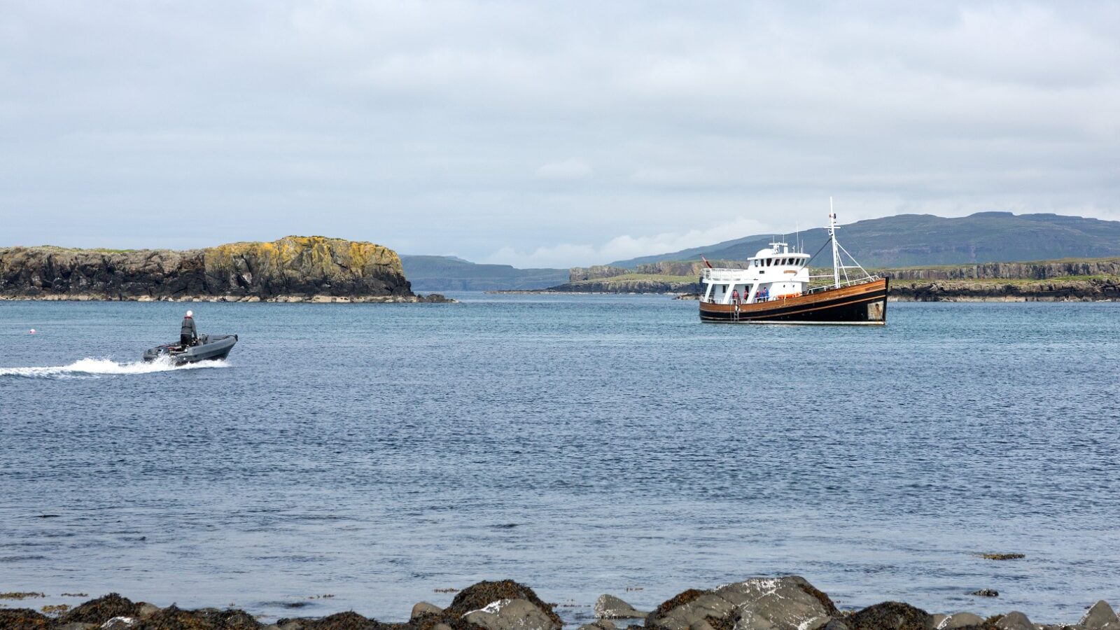 Two boats navigate calm waters near rocky shores and distant islands under a cloudy sky. The scene, reminiscent of an Argyll Cruising adventure, captures a serene maritime landscape.