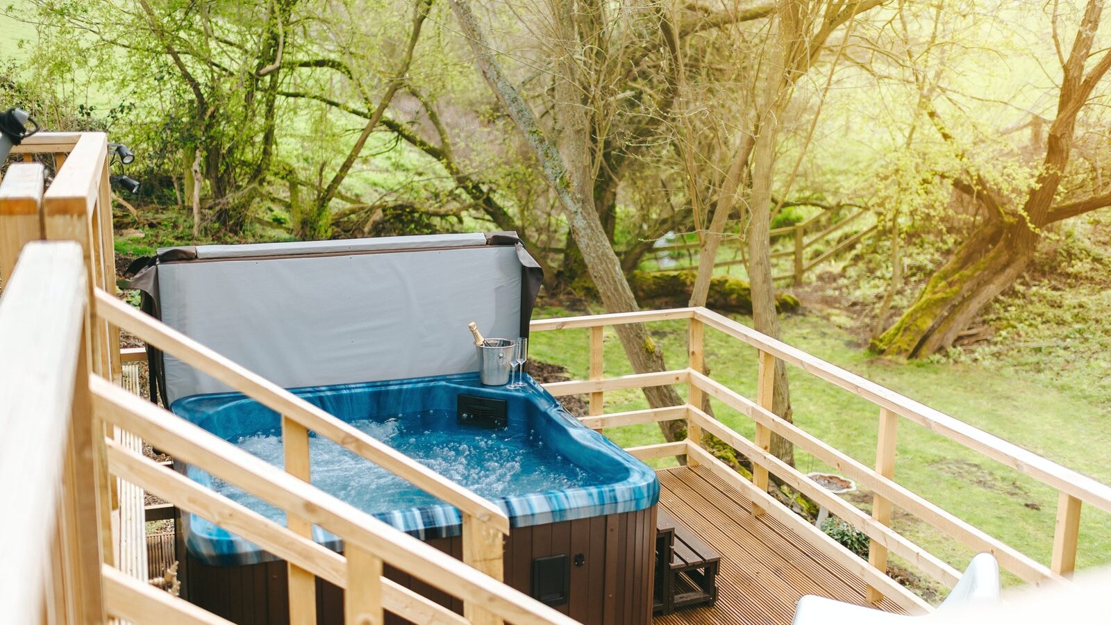 Nestled in the serene embrace of Bobs Lodge, an outdoor hot tub sits on a wooden deck, surrounded by lush green trees and grass.