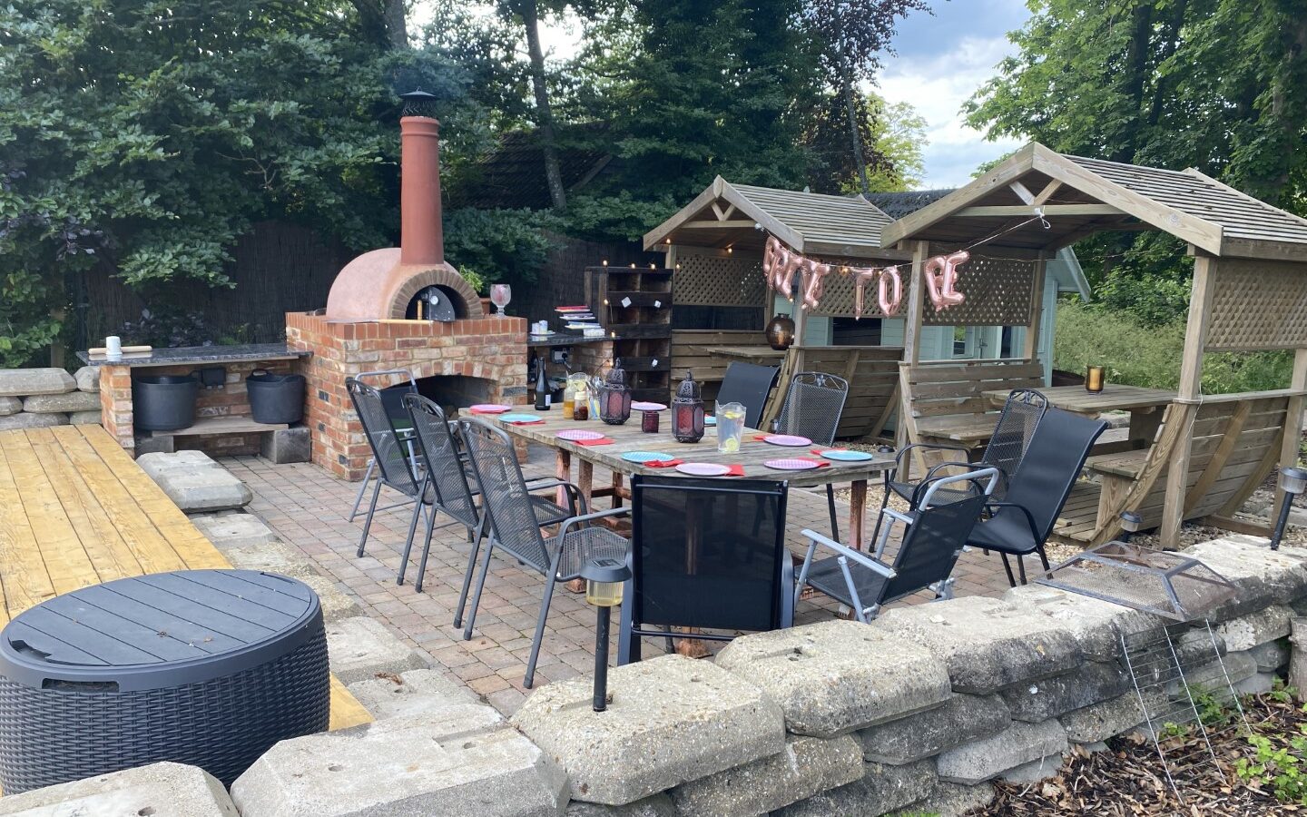 Nestled in a wooded backyard setting, the outdoor patio features a pizza oven, a long table set for dining, and chairs. With the charm of a rustic Chilterns View safari tent nearby, this serene space invites you to dine amidst nature's embrace.