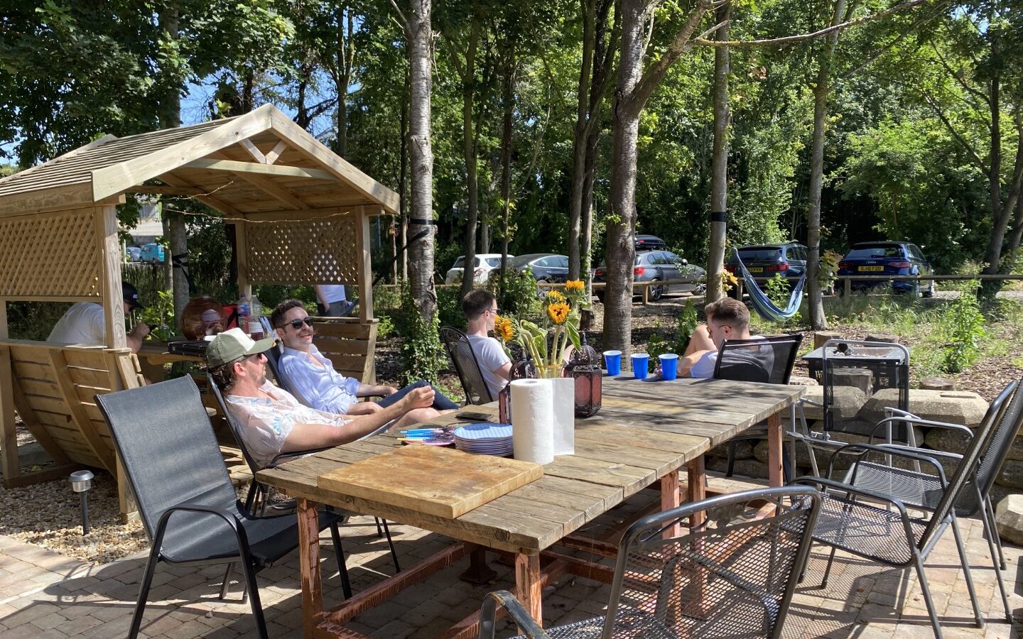 People sitting at outdoor tables with drinks and snacks on a sunny day, surrounded by trees and parked cars, enjoy the Chilterns View. It feels like a glamping retreat without stepping inside a safari tent.