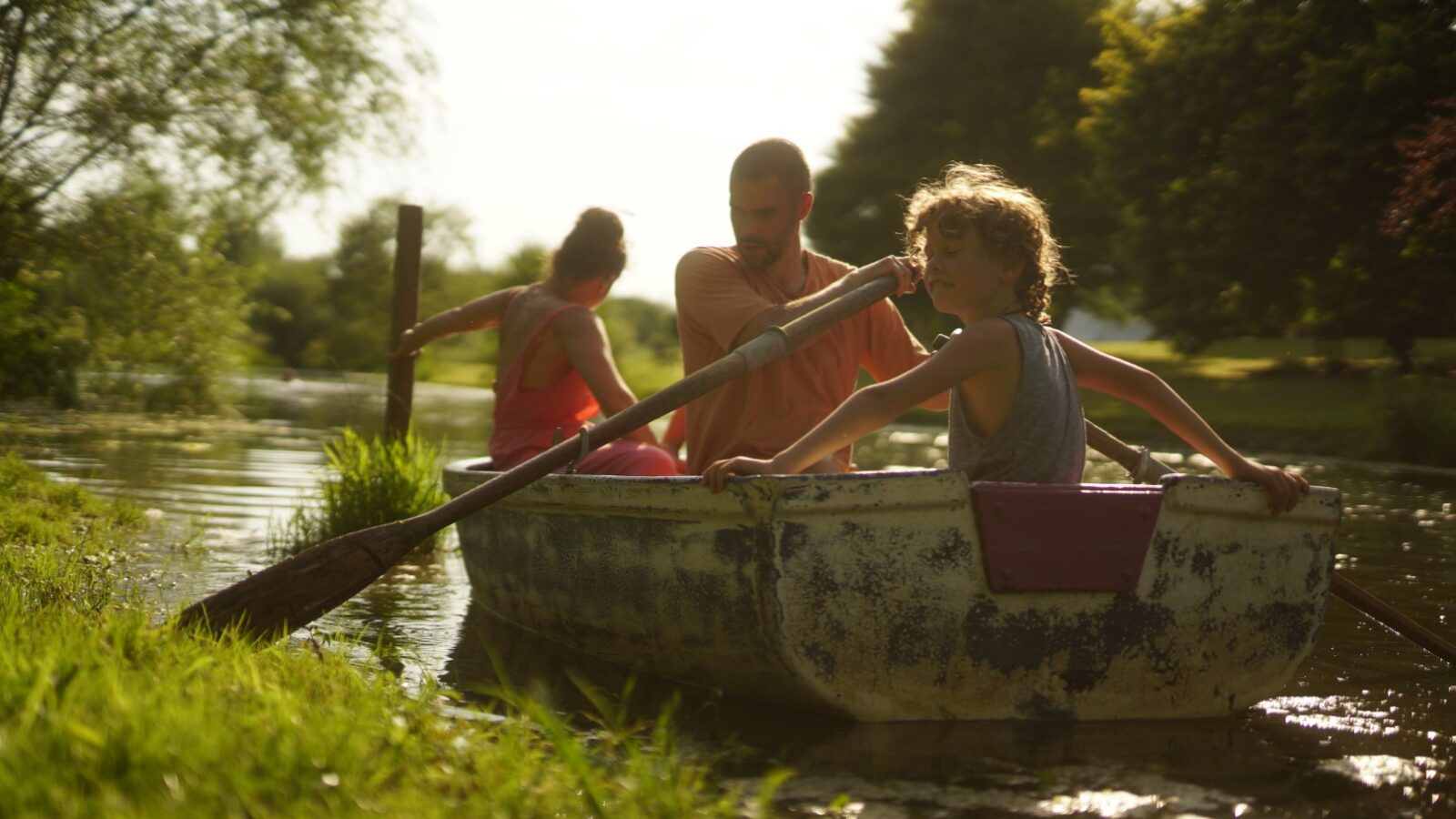 A family in a small boat paddles along a narrow, tree-lined river at West Lexham, enjoying the sunny day before returning to their cozy bell tents for a perfect glamping experience.