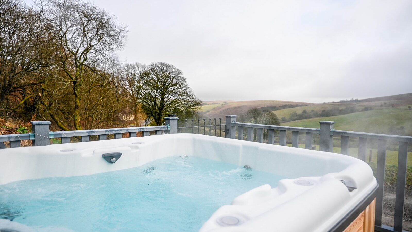 Enjoy a steaming hot tub on the wooden deck of your Beacon View Pods accommodation, with a stunning vista of rolling hills under an overcast sky. Experience glamping at its finest, where every moment is wrapped in tranquility and natural beauty.