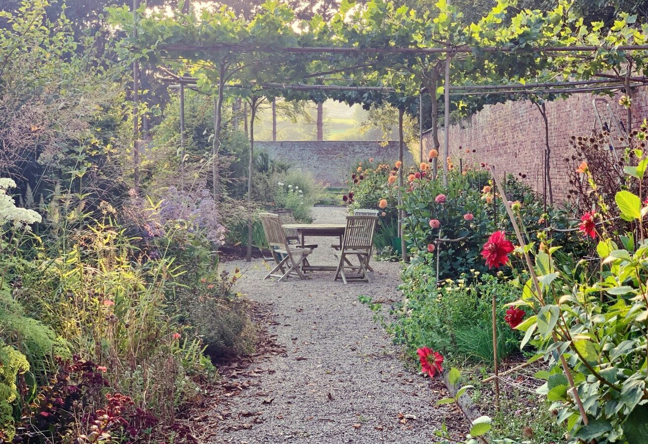 A garden path leads to a wooden table and chairs under a vine-covered pergola, offering a serene retreat surrounded by colorful flowers and lush greenery.