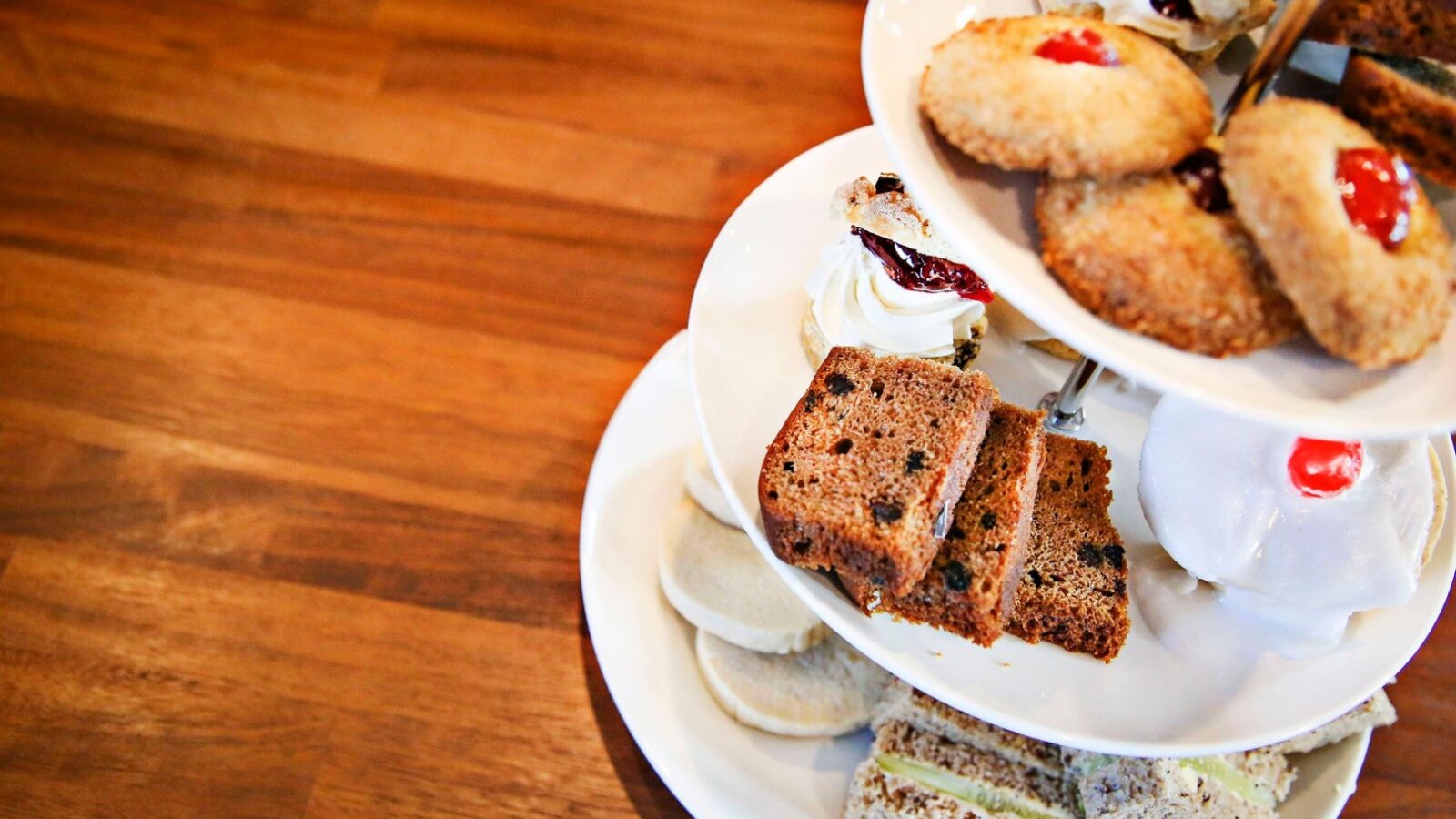 A multi-tiered stand displaying an assortment of pastries and sandwiches, reminiscent of a delightful Argyll Cruising afternoon tea. The top tier holds cookies and scones, the middle has slices of fruitcake, and the bottom features finger sandwiches, all set on a wooden table.