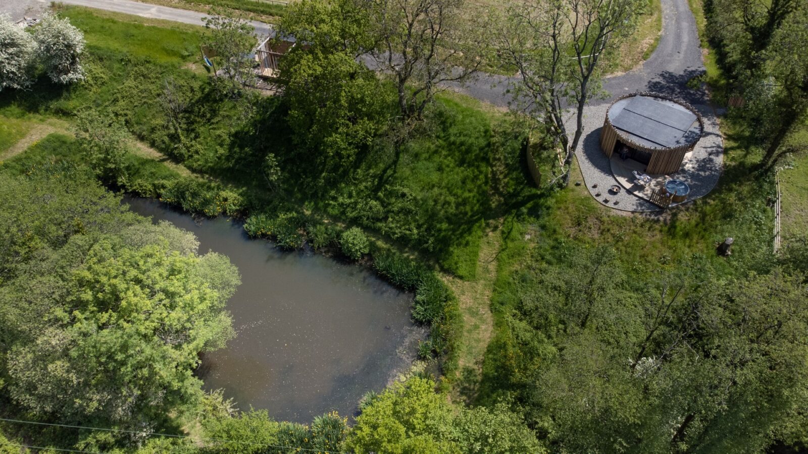 An aerial view captures Pond View Lodges, a circular wooden cabin with a patio set, nestled among lush green trees and grass. Nearby, a serene pond complements the idyllic setting while a gravel path winds its way to the cozy cabin.