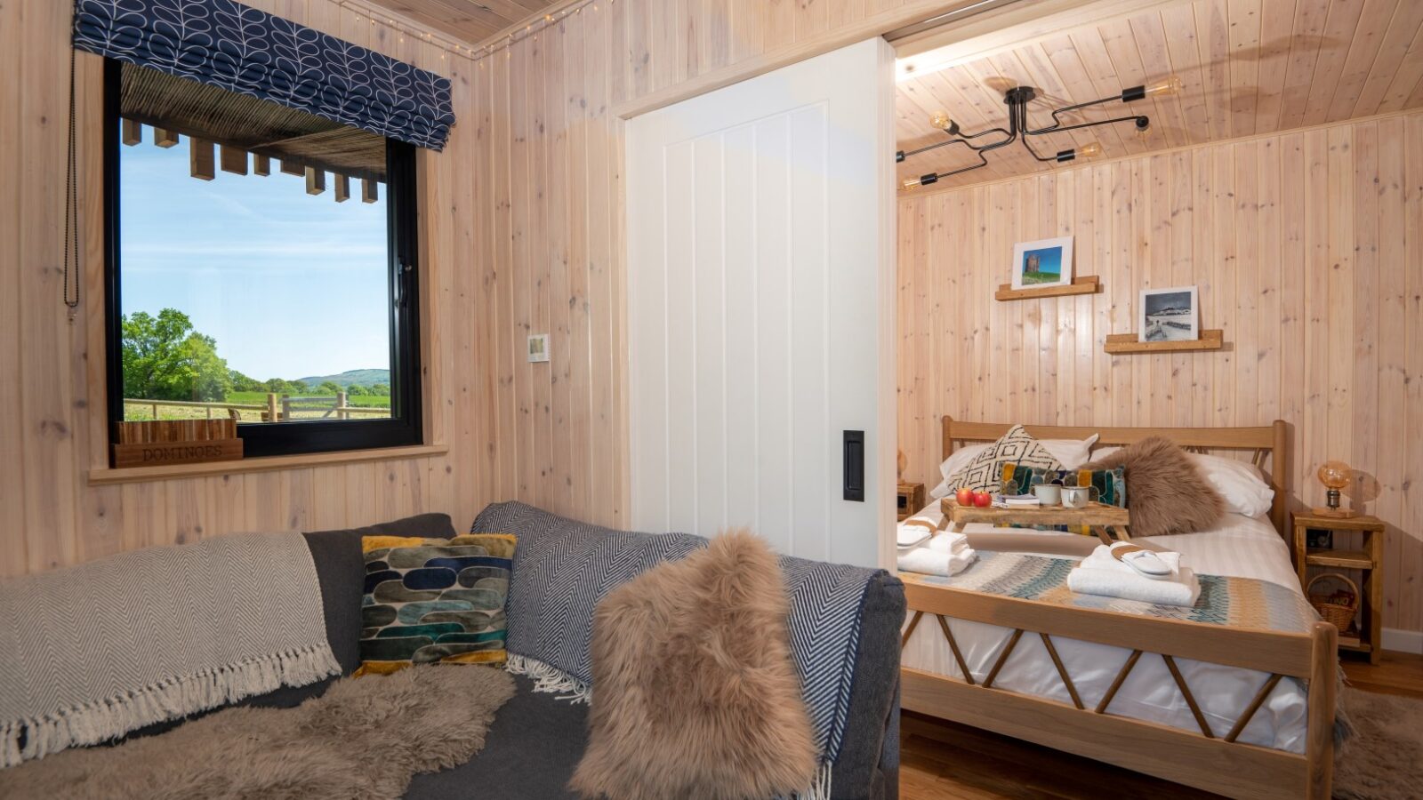 A cozy wooden cabin interior features a gray sofa with cushions and a fur throw, perfectly situated next to a window boasting a scenic pond view. The bedroom has a sliding door, neatly made bed with folded towels, and wooden shelves adorned with decor items, echoing the charm of Pond View Lodges.